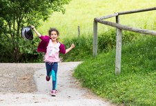Fröhliches Mädchen läuft mit Reithelm in der Hand einen Weg entlang beim Familienhotel Ponyhof in Kärnten.