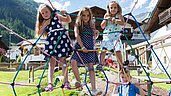 Drei Kinder spielen im Sommer auf dem Outdoor-Spielplatz des Familienhotels Alpenhotel Kindl in Tirol. Auf dem Outdoor Spielplatz hat man einen herrlichen Blick auf die umliegenden Berge.