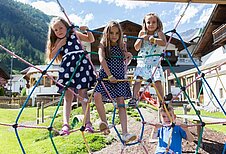 Drei Kinder spielen im Sommer auf dem Outdoor-Spielplatz des Familienhotels Alpenhotel Kindl in Tirol. Auf dem Outdoor Spielplatz hat man einen herrlichen Blick auf die umliegenden Berge.