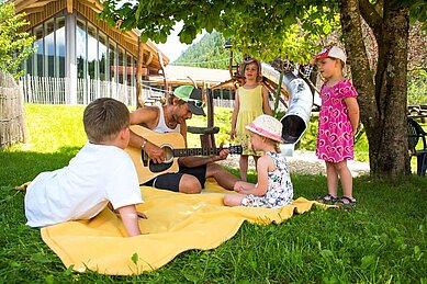 Ein Kinderbetreuer spielt Gitarre und die Kinder hören aufmerksam zu im Garten des Familienhotels Spa- & Familien-Resort Krone im Allgäu.