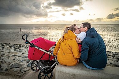 Eine glückliche Familie genießt einen entspannten Spaziergang auf dem Deich an der Nordsee, wobei die Eltern einen Kinderwagen schieben und ein malerischer Sonnenuntergang über dem ruhigen Meer den Hintergrund bildet.