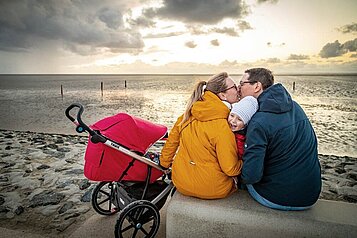 Eine glückliche Familie genießt einen entspannten Spaziergang auf dem Deich an der Nordsee, wobei die Eltern einen Kinderwagen schieben und ein malerischer Sonnenuntergang über dem ruhigen Meer den Hintergrund bildet.