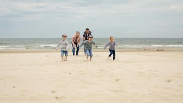 Familie mit drei Kindern rennt lachend am Sandstrand entlang, während das Meer im Hintergrund sanft an die Küste brandet.