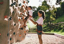 Ein Mädchen steht vor dem Kletterfels im Outoorgelände vom Familienhotel Post Family Resort im Salzburger Land.