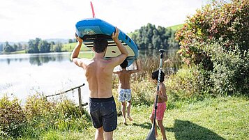 Vater mit seinen beiden Kindern am Seeufer mit einem SUP auf den Schultern.