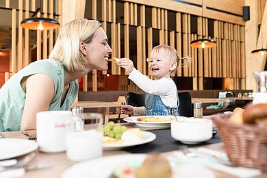 Mutter und Kind essen gemeinsam im Familienhotel. Kind füttert Mutter mit einem Stück Käse. 