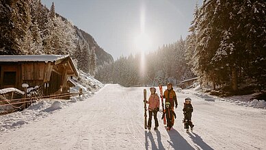 Eine Familie bereitet sich auf dem Weg zum Skifahren an einem sonnigen Wintertag am Hotel Habachklause im Salzburger Land vor, umgeben von schneebedeckten Bäumen.