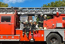 Kinder sitzen auf Oldtimer Feuerwehrauto in Feuerwehranzügen und zeigen nach links oben im Familienhotel Ulrichshof im Bayerischen Wald