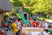 Familie sitzt auf der Terrasse des Familienhotels Sonne Bezau und genießt ein Eis, während das andere Kind die Rutsche hinunter rutscht.