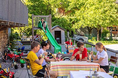 Familie sitzt auf der Terrasse des Familienhotels Sonne Bezau und genießt ein Eis, während das andere Kind die Rutsche hinunter rutscht.