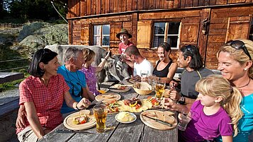 Große Familie sitzt auf einer Almhütte und macht Brotzeit nach einer Wanderung.