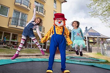 Das Familotel-Maskottchen Happy mit zwei Kindern auf dem Outdoor-Trampolin.