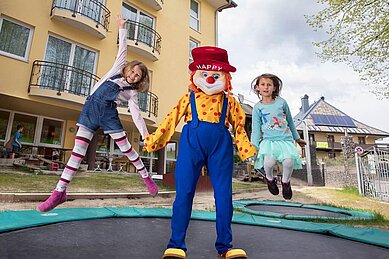 Das Familotel-Maskottchen Happy mit zwei Kindern auf dem Outdoor-Trampolin.