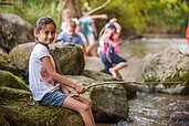 Mädchen sitzt mit einer selbst gebastelten Angel aus Holz am Bach und versucht zu fischen.