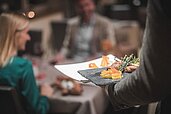 Ein Mitarbeiter bringt zwei Teller mit leckerem Essen an den Tisch im Familienhotel Alpenhof Dolomit Family.