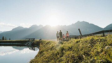 Familie wandert entlang des Sees im Stubaital, im Hintergrund ist das Alpenpanorama zu sehen.