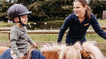 Kind sitzt auf dem Reitplatz des Familienhotels Landhaus Averbeck in der Lüneburger Heide auf einem Pony und wird von einer Reitlehrerin geführt.