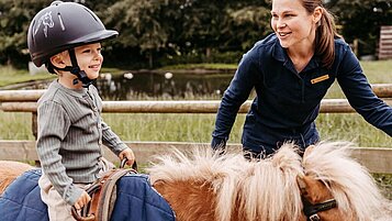 Kind sitzt auf dem Reitplatz des Familienhotels Landhaus Averbeck in der Lüneburger Heide auf einem Pony und wird von einer Reitlehrerin geführt.