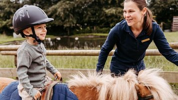 Kind sitzt auf dem Reitplatz des Familienhotels Landhaus Averbeck in der Lüneburger Heide auf einem Pony und wird von einer Reitlehrerin geführt.