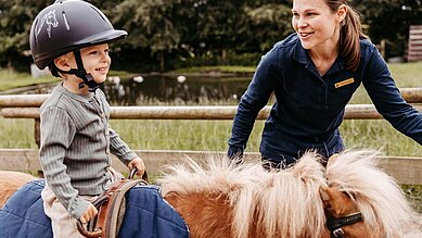 Kind sitzt auf dem Reitplatz des Familienhotels Landhaus Averbeck in der Lüneburger Heide auf einem Pony und wird von einer Reitlehrerin geführt.