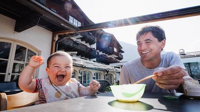 Ein lachendes Baby sitzt mit einem Erwachsenen an einem Tisch auf der Sonnenterrasse des Familienhotels Tirolerhof beim Füttern.