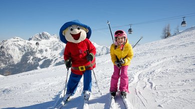 Ein glückliches Kind fährt im Familienhotel Ramsi in Kärnten mit dem Maskottchen auf der Skipiste, umgeben von einer beeindruckenden Bergkulisse