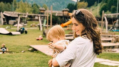 Junge Mutter hält ihr Kleinkind auf dem Arm und blickt lächelnd auf den weitläufigen Outdoor-Spielplatz im Alpura Retreat mit Bergpanorama im Hintergrund.