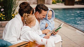 Familie mit zwei Kindern sitzt im Bademantel am Außenpool des Familienhotels Landhaus Averbeck in der Lüneburger Heide. Der Vater liest ein Buch vor.