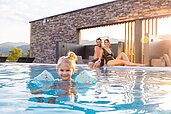 Mädchen schwimmt mit Schwimmflügeln im Außenpool des Familienhotels Schreinerhof, die Eltern sitzen am Beckenrand und passen auf,.