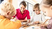 Eine Kinderbetreuerin sitzt mit drei Kindern am Tisch und malt im Familienhotel Allgäuer Berghof.