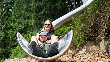 Mutter rutscht gemeinsam mit ihrem Kind eine Röhrenrutsche im Wald hinunter.