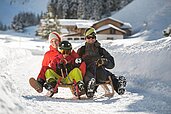 Dreiköpfige Familie beim Schlittenfahren auf der familienfreundlichen Rodelbahn im Familienurlaub in Tirol, umgeben von einer verschneiten Winterlandschaft.