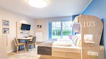 Familienfreundliches Zimmer im Familienhotel Deichkrone mit einem hellen und einladenden Interieur, ausgestattet mit einem großen Bett mit Spielhaus-Design, gemütlichen Sitzgelegenheiten und direktem Zugang zur grünen Außenterrasse, ideal für eine entspannende Familienauszeit an der Nordsee.