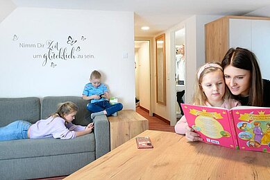 Mutter sitzt mit ihrer Tochter im Familienzimmer am Tisch und liest ein Buch. Zwei andere Kinder liegen auf der Couch und der Vater macht sich im Badezimmer fertig.