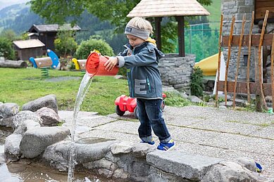 Ein Kind schüttet einem Eimer Wasser in ein Wasserbecken am Wasserspielplatz im Familienhotel Alphotel