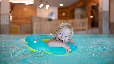 Das kleine Kind treibt in seinem Schwimmring im Indoor-Schwimmbad des Familienhotels Landhaus zur Ohe im Bayerischen Wald umher und lächelt dabei über beide Ohren.