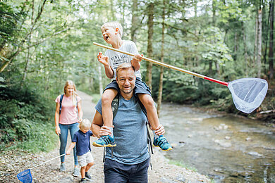 Familie mit zwei Kindern wandert im Wald am Fluss entlang. Die Kinder haben Kescher in der Hand.