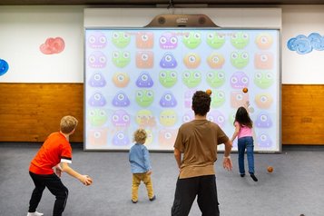 Kinder spielen begeistert ein interaktives Ballspiel vor einer großen digitalen Spielwand im Spielzimmer des Hotels Feldberger Hof.