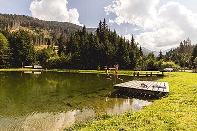 Zwei Kinder springen vom Steg in einen Natur-Badeteich am Almfamilyhotel Scherer in Tirol.