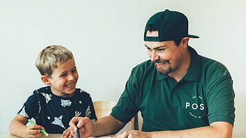 Ein Kinderbetreuer und ein Junge sitzen an einem Tisch mit Malsachen im Familienhotel Post Family Resort im Salzburger Land.