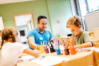 Betreuer sitzt mit zwei Kindern im Happy-Club und malt mit ihnen ein Bild im Familienhotel Das Bayrischzell in Oberbayern.