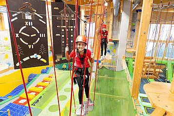Ein lächelndes Kind in einem roten Helm und Sicherheitsgurt genießt das Klettern in einem Indoor-Spielplatz mit Seilen und Kletterwänden im Hotel Feldberger Hof.