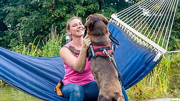 Mutter sitzt auf einer Hängematte im Garten des Familienhotels Engel und spielt mit ihrem Hund.