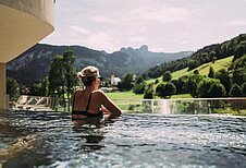 Eine Frau steht im Infinitypool und genießt die Aussicht auf die umliegenden Berge im Familienhotel Post Family Resort im Salzburger Land.