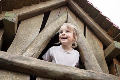 Ein fröhliches Kind schaut aus dem Fenster eines Holzkletterturms am Spielplatz eines Familotel.