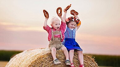 Kinder sitzen in Tracht auf einem Heuballen und halten Brezen in die Luft im Familienhotel Bavaria im Allgäu.