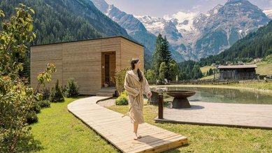 Ein Steg führt um einen kleinen Naturteich zur außenliegenden Sauna, im Hintergrund Berge beim Hotel Bella Vista in Trafoi in Südtirol.