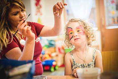 Kinderbetreuerin im Happy-Club des Familienhotels Landgut Furtherwirt bemalt das Gesicht eines Mädchens mit bunten Farben.