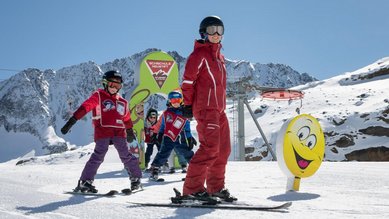 Kinder lernen mit einem Skilehrer in der Skischule im Familienhotel Alpenhotel Kindl im Stubaital das Skifahren in der Sonne vor beeindruckender Bergkulisse.
