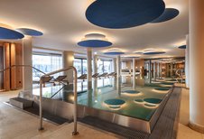 Großzügiger Innenpool im Zauchenseehof mit modernen Deckenelementen in Blautönen, stimmungsvoller Beleuchtung und Liegen entlang der Panoramafenster.